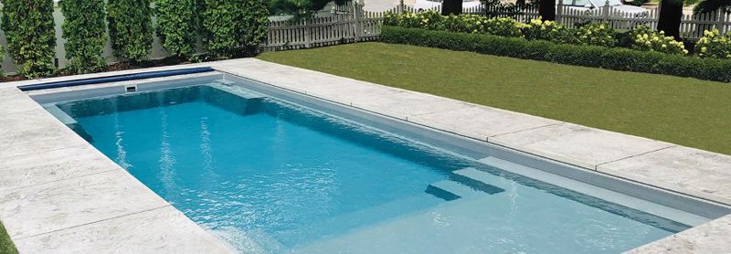 A rectangular fiberglass pool with a corner step, set in a paved patio beside a clipped lawn.