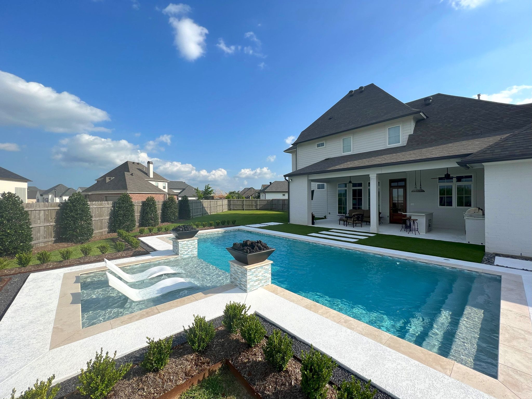 A rectangular pool with two in-water loungers on a shallow ledge and two unlit fire bowls on tiled plinths, in the back yard of a two-storey house.