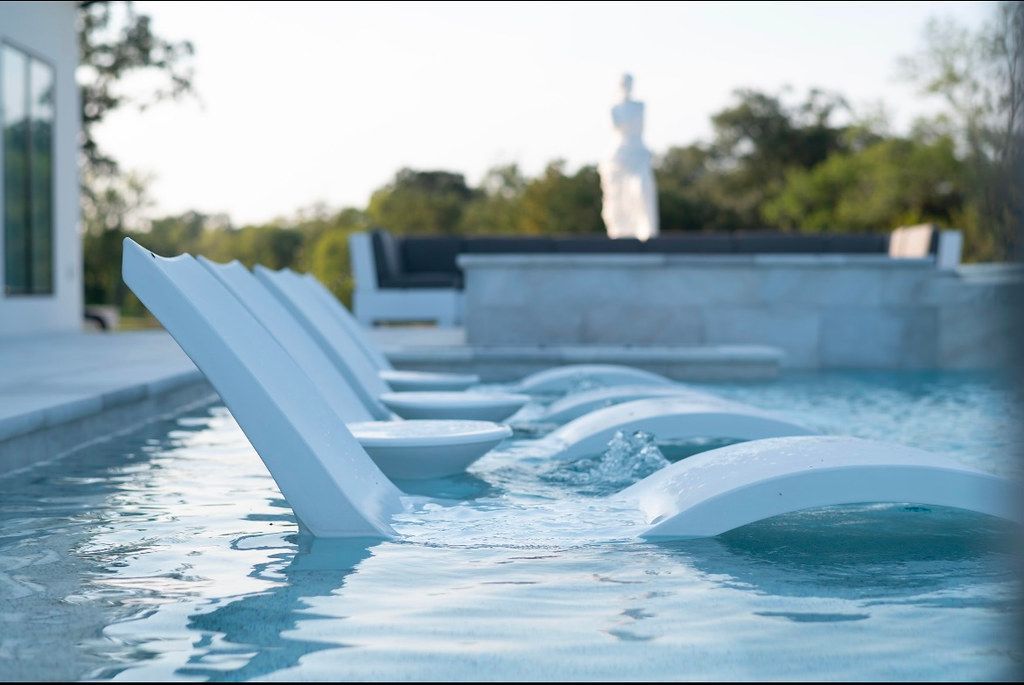 In-water lounge chairs on a shallow ledge of a gunite pool, with a sculpture and trees beyond.