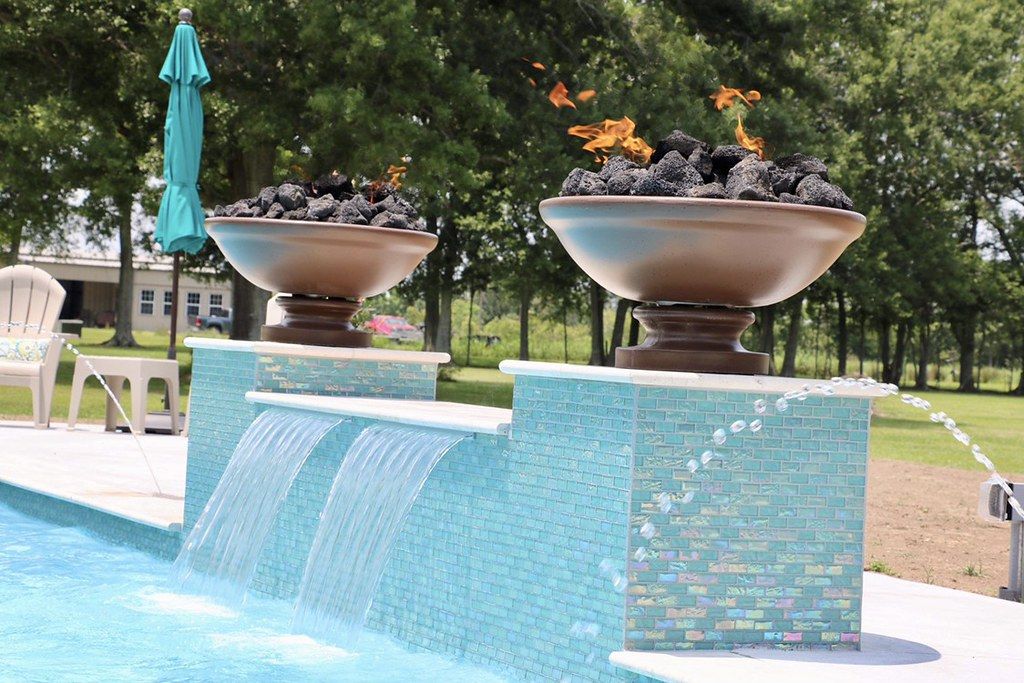 Two lit fire bowls on tiled plinths above two sheet waterfalls and a single arcing laminar jet, all spilling into a gunite pool.