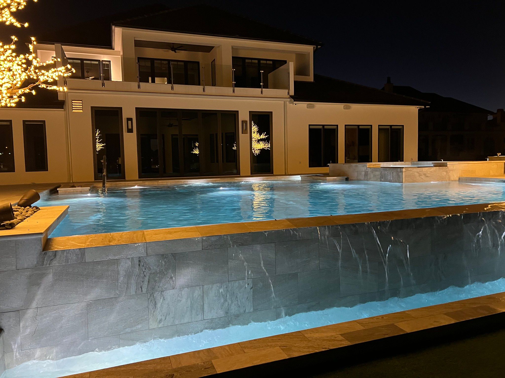 A raised gunite pool at night, water sheeting over a stone-clad wall and lit from within, in front of a modern house.