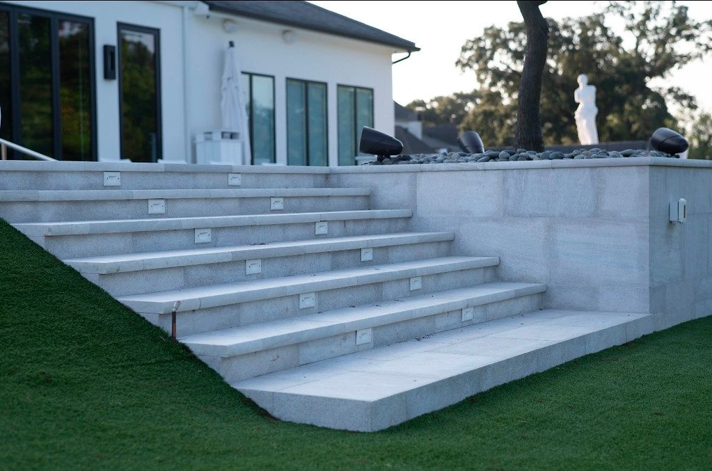 Wide stone steps with recessed lights leading up to a raised pool deck beside a white house.