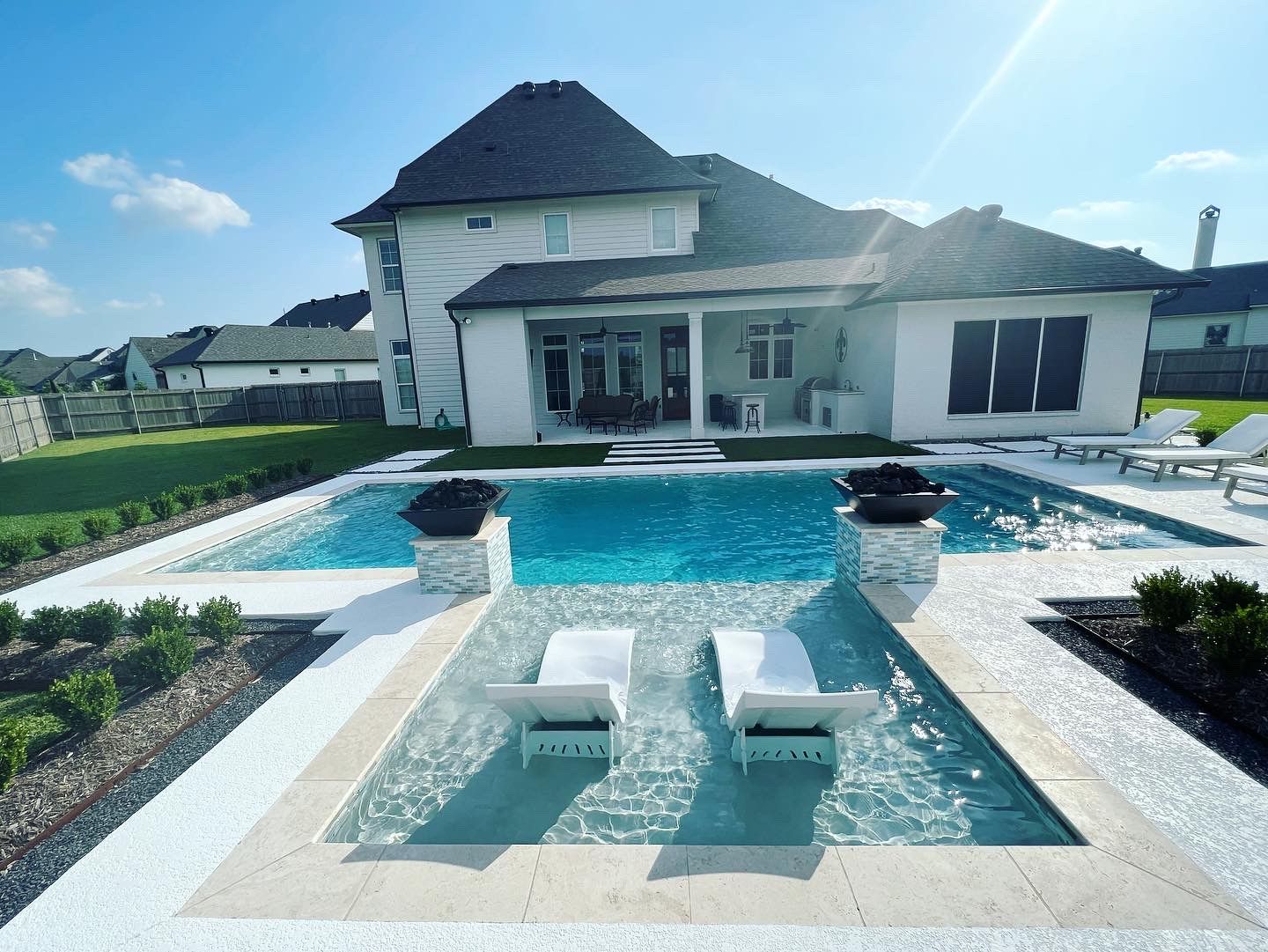 A gunite pool with a broad tanning ledge holding two in-water loungers, behind a large white house.